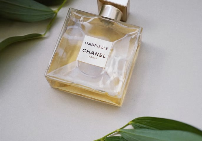 A glass bottle of Gabrielle Chanel perfume sits on a white table surrounded by green leaves