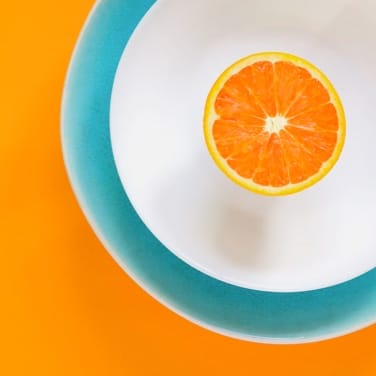 Half an orange balanced peel down on a white plate with a blue border, all on an orange table.